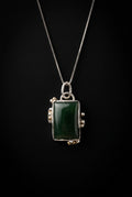 A silver-toned necklace with a rectangular dark green stone pendant, displayed on a dark surface.