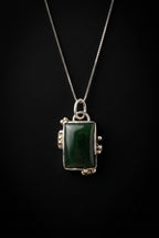 A silver-toned necklace with a rectangular dark green stone pendant, displayed on a dark surface.