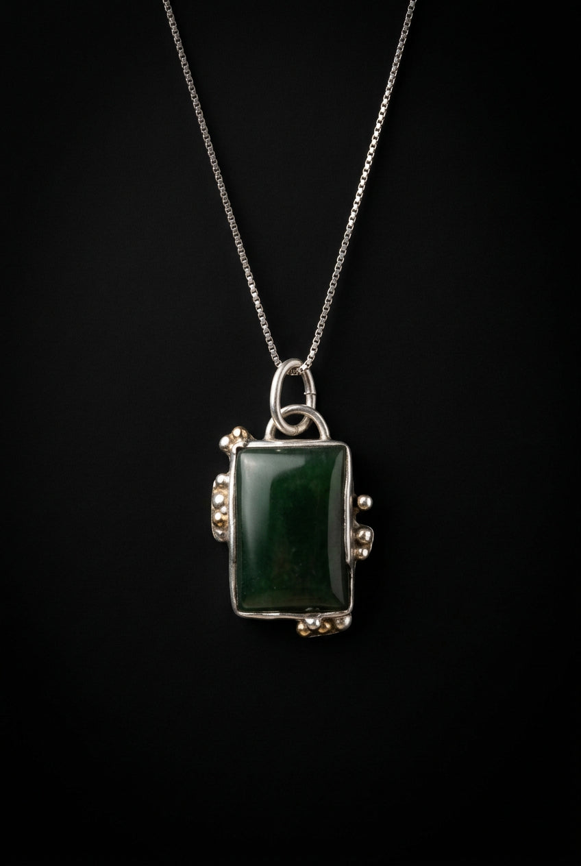 A silver-toned necklace with a rectangular dark green stone pendant, displayed on a dark surface.