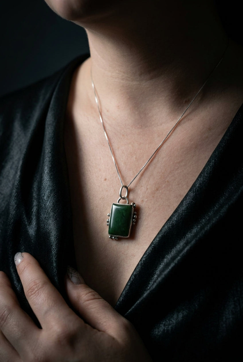 A close-up of a person wearing a rectangular green pendant necklace with a rose gold-toned frame and chain, styled with a black satin top against a dark background.