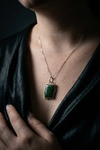 A close-up of a person wearing a rectangular green pendant necklace with a rose gold-toned frame and chain, styled with a black satin top against a dark background.