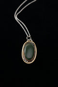 An oval pendant necklace featuring a dark green cabochon stone set in a double-layered gold frame with small silver accents, suspended from a silver chain against a dark background.