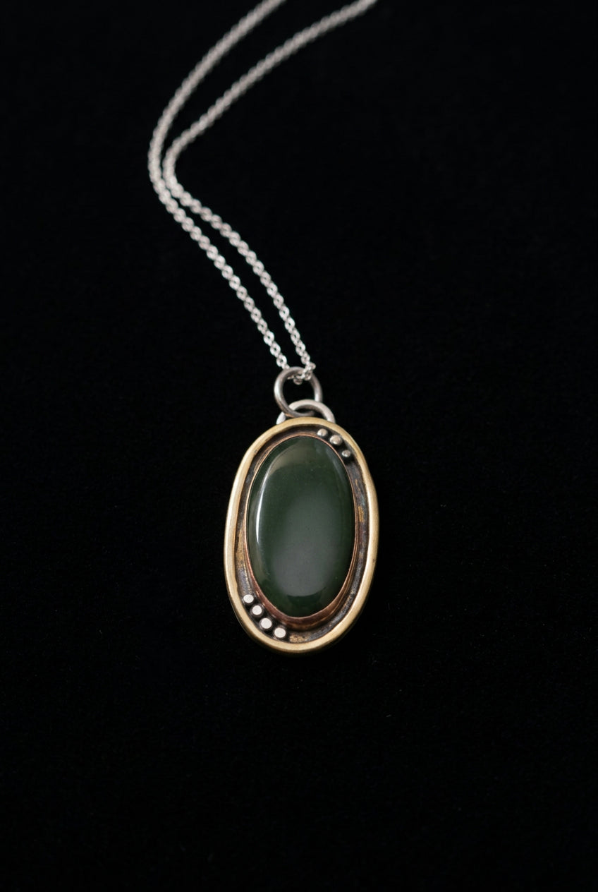 An oval pendant necklace featuring a dark green cabochon stone set in a double-layered gold frame with small silver accents, suspended from a silver chain against a dark background.