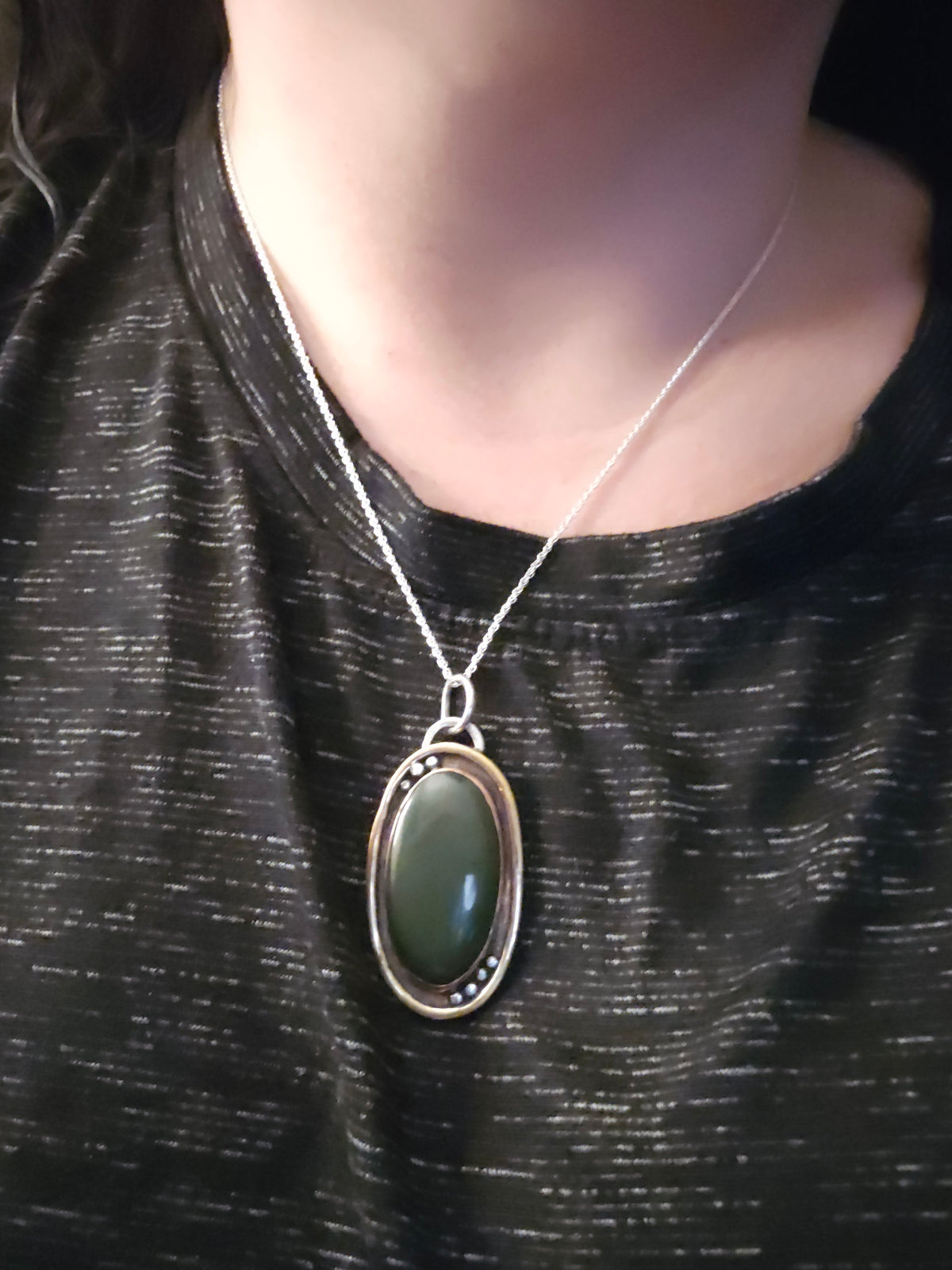 A close-up of a person wearing a silver-toned oval pendant necklace with a dark green stone, shown against a black textured top.