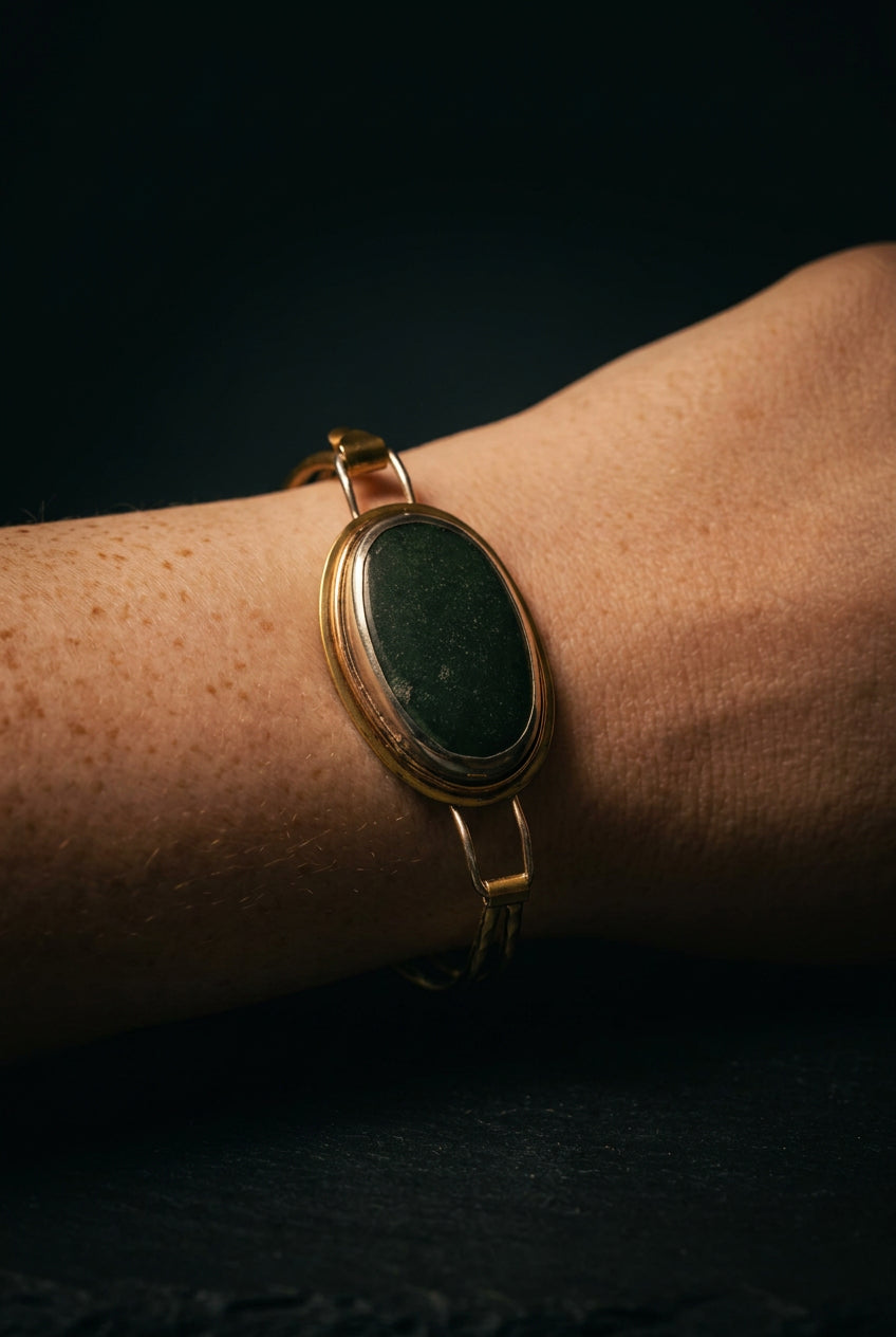 A gold-toned oval bezel bracelet with a dark green stone, worn on a person's wrist alongside a thinner gold bangle.