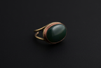 A vintage-style ring featuring a large, smooth oval dark green stone in a warm bronze-toned metal setting, shown on a dark black background.