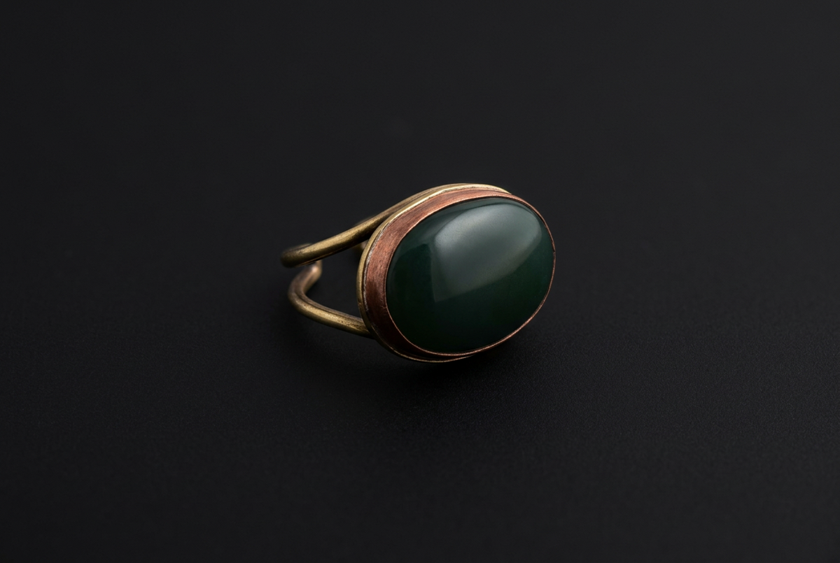 A vintage-style ring featuring a large, smooth oval dark green stone in a warm bronze-toned metal setting, shown on a dark black background.