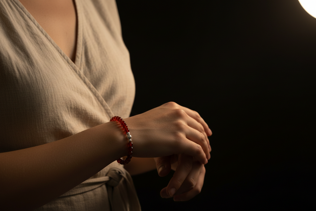 A close-up lifestyle shot of a person wearing a red beaded bracelet on their wrist, wearing a light beige wrap dress against a dark background.