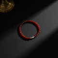 A beaded stretch bracelet made of translucent red-orange beads with a small silver accent bead, photographed on a dark surface with dramatic sunlight and shadows.
