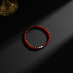 A beaded stretch bracelet made of translucent red-orange beads with a small silver accent bead, photographed on a dark surface with dramatic sunlight and shadows.