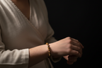 A close-up of a person wearing a light beige linen wrap top and a gold beaded bracelet on their wrist, set against a solid black background.