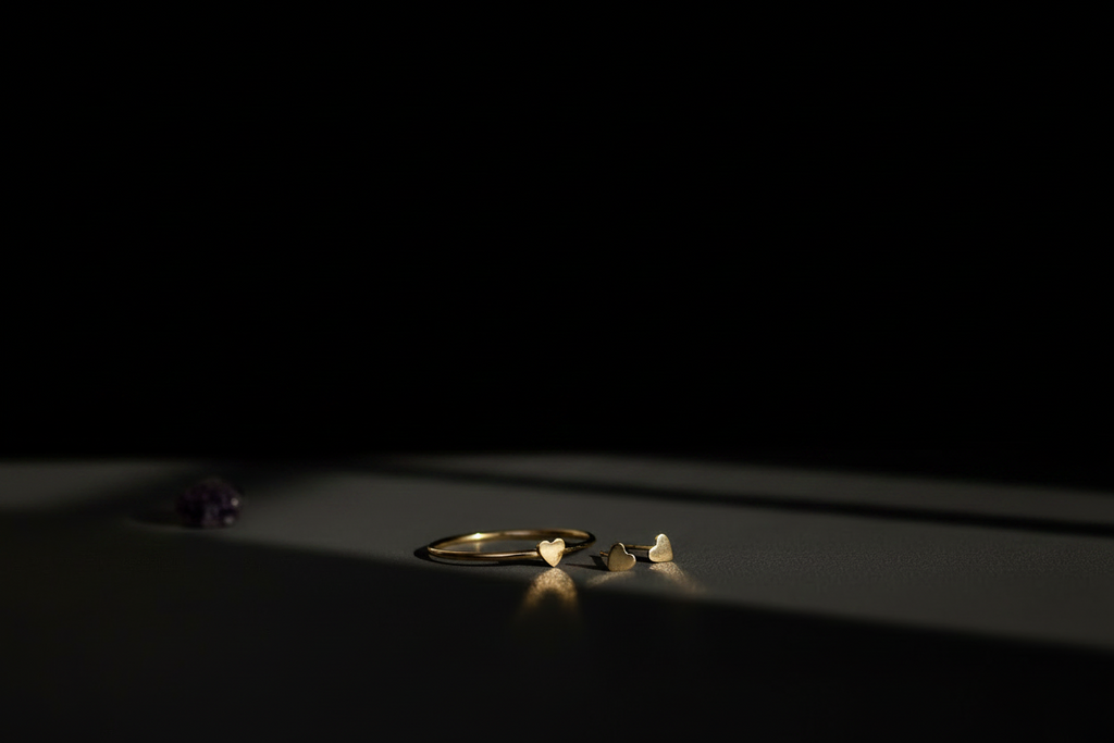 A minimalist jewelry display featuring a gold ring and matching small gold earrings on a dark surface, with dramatic spotlighting against a solid black background.