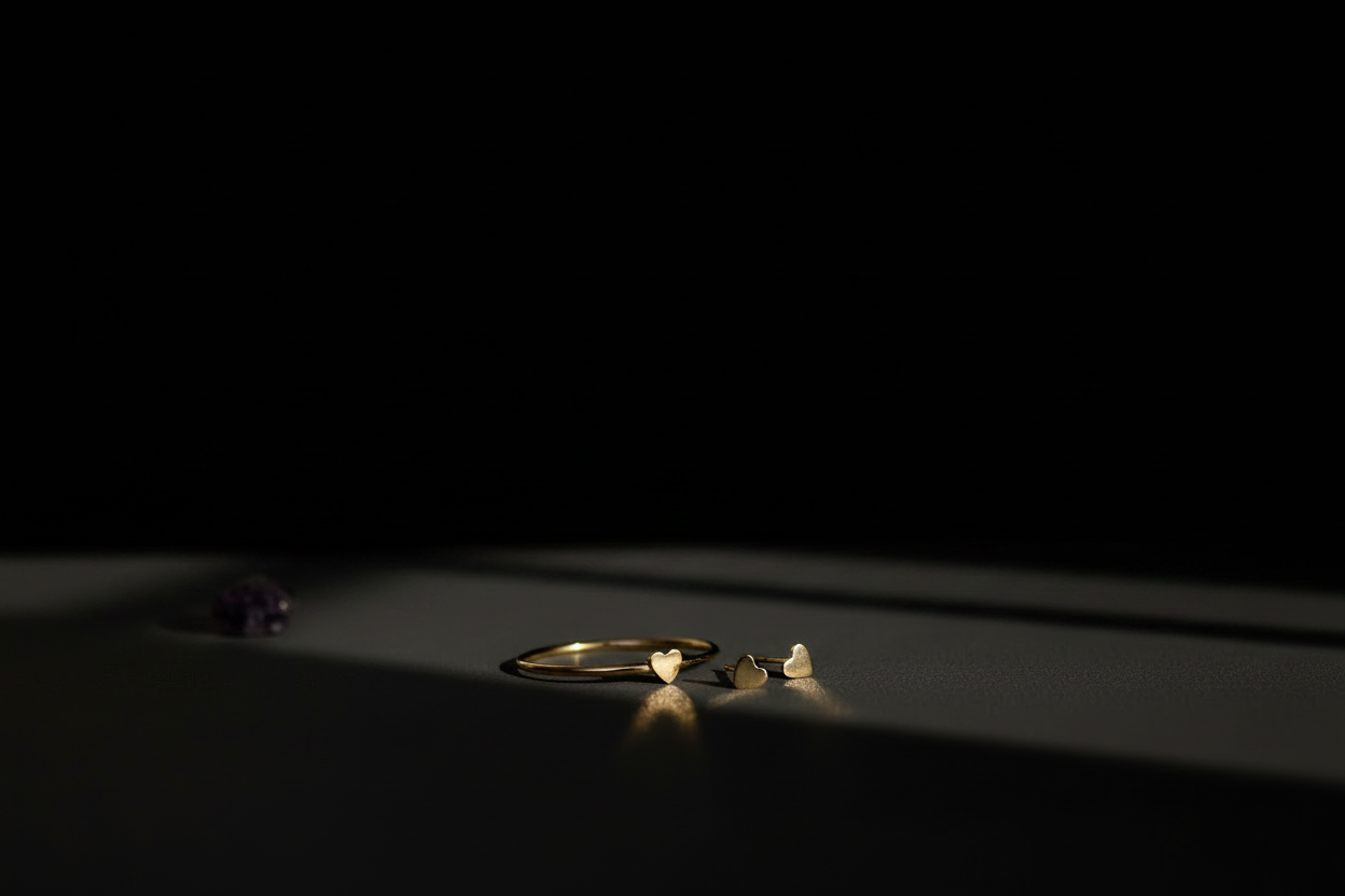 A minimalist jewelry display featuring a gold ring and matching small gold earrings on a dark surface, with dramatic spotlighting against a solid black background.
