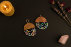 A pair of handmade bohemian earrings featuring engraved wooden semi-circles and multi-strand beaded fringe in vibrant colors, displayed on a dark background with a lit candle, lavender, and a pink crystal.