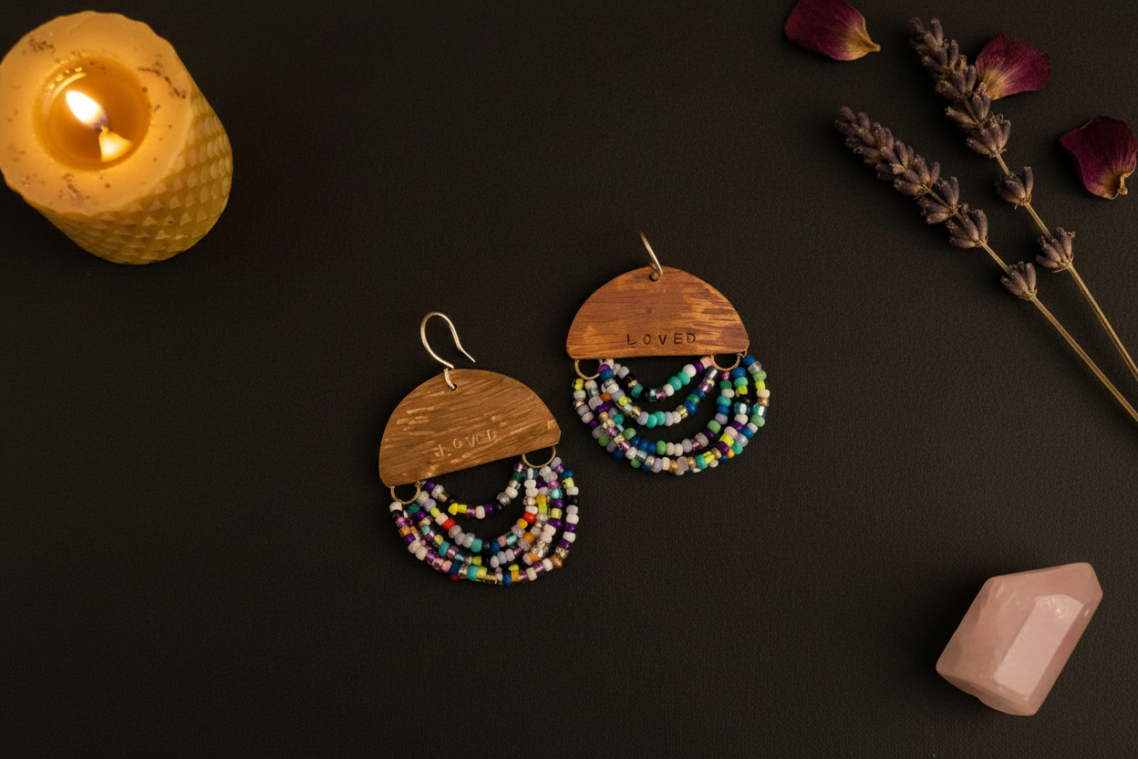 A pair of handmade bohemian earrings featuring engraved wooden semi-circles and multi-strand beaded fringe in vibrant colors, displayed on a dark background with a lit candle, lavender, and a pink crystal.