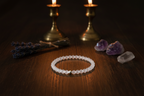 A beaded bracelet made of small translucent white beads with a tiny gold accent, displayed on a dark wooden surface with candles, lavender, and crystals in the background.
