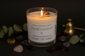 A lit candle in a clear glass jar with a white label reading 'Manifestation' and 'Frosted Cinnamon Bun,' styled on a dark surface with crystals, green leaves, and a brass bell.