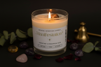 A lit candle in a clear glass jar with a white label reading 'Manifestation' and 'Frosted Cinnamon Bun,' styled on a dark surface with crystals, green leaves, and a brass bell.