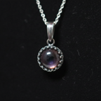 A silver pendant necklace featuring a round iridescent purple gemstone surrounded by a silver beaded halo, hanging from a silver chain against a black background.