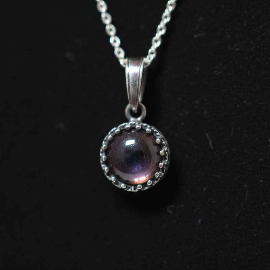 A silver pendant necklace featuring a round iridescent purple gemstone surrounded by a silver beaded halo, hanging from a silver chain against a black background.