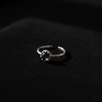 A silver-toned open band ring with a textured, organic-looking setting holding a round, dark iridescent gemstone, resting on a dark textured surface.