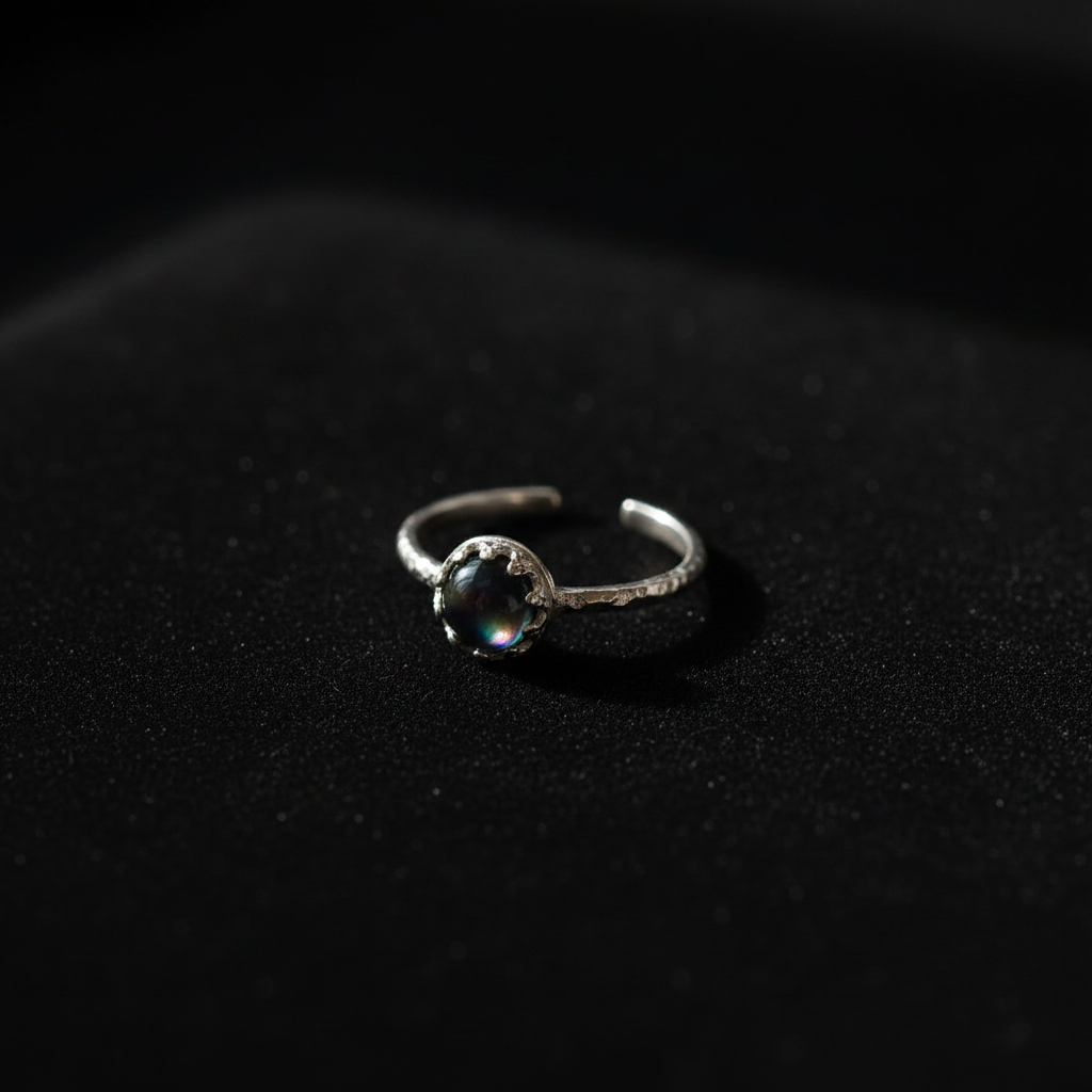 A silver-toned open band ring with a textured, organic-looking setting holding a round, dark iridescent gemstone, resting on a dark textured surface.
