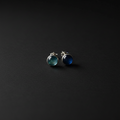 A pair of silver stud earrings with round cabochon gemstones, one greenish-blue and one deep blue, displayed on a dark textured background.