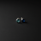 A pair of silver stud earrings with round cabochon gemstones, one greenish-blue and one deep blue, displayed on a dark textured background.