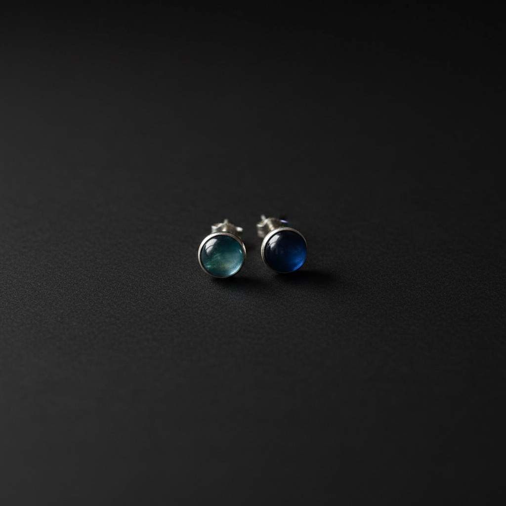 A pair of silver stud earrings with round cabochon gemstones, one greenish-blue and one deep blue, displayed on a dark textured background.