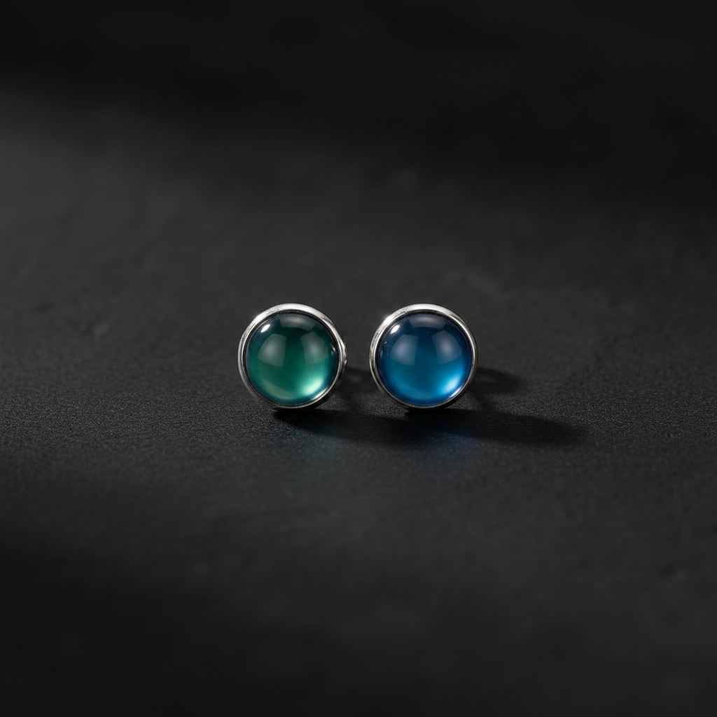 A pair of silver stud earrings with round cabochon gemstones, one greenish-blue and one blue, displayed on a dark textured surface.