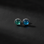 A pair of silver stud earrings with round cabochon gemstones, one greenish-blue and one blue, displayed on a dark textured surface.