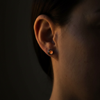 A close-up side profile of a person's ear wearing a small round stud earring with a dark metallic setting and a warm amber-orange center stone, set against a dark background.