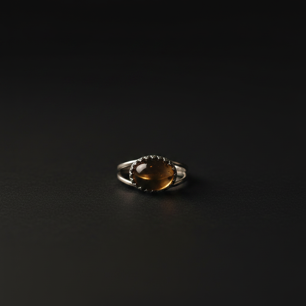 A silver ring featuring an oval amber-brown cabochon gemstone surrounded by a dark beaded halo, set against a dark black background.