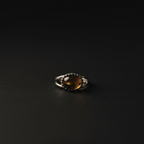 A silver ring featuring an oval amber-brown cabochon gemstone surrounded by a dark beaded halo, set against a dark black background.