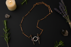 A copper-toned chain necklace with a unique pendant featuring two large silver nails wrapped in dark beads and connected by a silver ring, displayed on a dark background with candles, rosemary, and lavender.