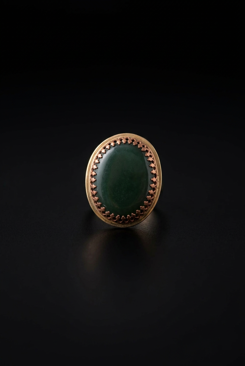 An oval ring featuring a smooth, polished dark green cabochon stone set in a textured  copper-toned bezel with a starburst border, photographed on a dark black background.