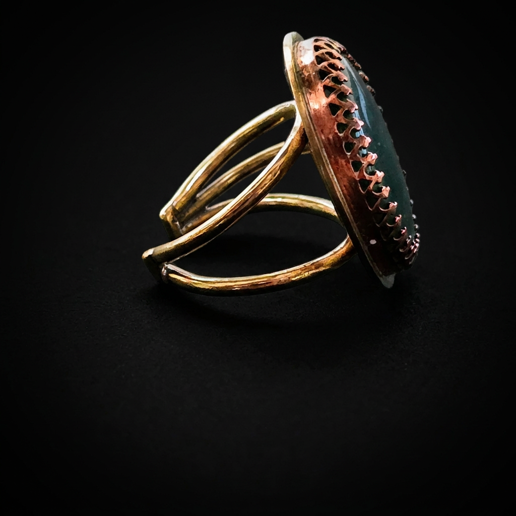 A rustic open-band ring featuring a large oval cabochon stone with a copper-toned textured border and a dark green center, set against a solid black background.