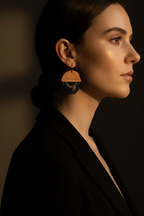 A woman in profile wearing a large geometric dangle earring with a warm tan top section and dark blue beaded fringe, photographed in dramatic warm lighting against a dark background.