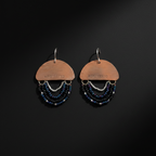 A pair of handmade dangle earrings on a dark black background. Each earring features a copper-colored metal semicircle stamped with the word 'PROTECTED' and hangs from silver hooks.