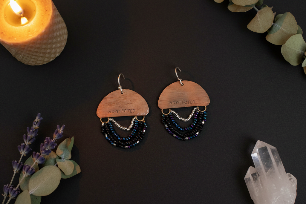 A pair of handmade dangle earrings displayed on a dark background with decorative props including a lit candle, lavender, eucalyptus leaves, and a crystal cluster. The earrings feature a wooden semicircle top engraved with 'THE JOY OF IT', a silver fishhook, and three cascading beaded fringe layers in black, silver, and dark blue.