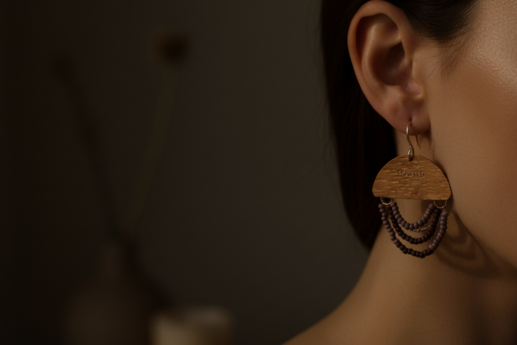 A close-up side view of a person's ear wearing a dangling earring. The earring has a light natural wood top with engraved text, three curved rows of dark beads, and a small gold hoop at the top.