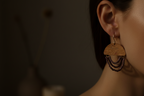 A close-up side view of a person's ear wearing a dangling earring. The earring has a light natural wood top with engraved text, three curved rows of dark beads, and a small gold hoop at the top.