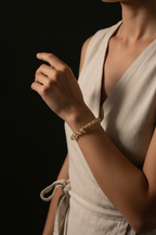 A close-up of a woman wearing a delicate gold beaded bracelet on her wrist, dressed in a light beige linen wrap dress against a solid black background.