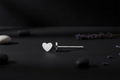 A single silver stud earring with a small, polished heart-shaped face, displayed on a dark textured surface with decorative stones and lavender sprigs.