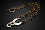 A handmade link chain necklace featuring alternating bronze and gold-toned metal rings, with a central silver crescent moon and disc pendant accented by small gold and purple details, displayed on a dark background.