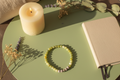 Beaded bracelet on a green surface with a lit candle and notebook in the background