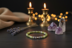 A beaded bracelet made of light green and gray beads with a small metallic accent, displayed on a dark wooden surface with candles, lavender, crystals, and warm string lights in the background.