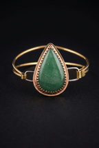 Handcrafted mixed-metal bangle bracelet featuring a teardrop green aventurine cabochon set in a scalloped copper crown bezel with double brass wire bands and clasp loops, on a dark moody background.