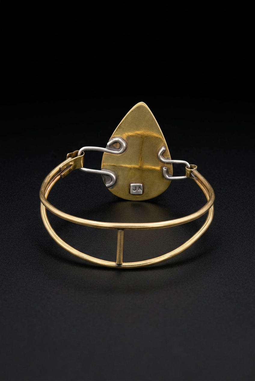 Inside back view of a handcrafted mixed-metal teardrop bangle bracelet displaying the patinated brass backplate, maker's hallmark stamp, and silver connector links on a dark surface.