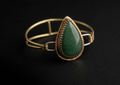Side profile view of a handcrafted mixed-metal bangle bracelet showcasing a teardrop green aventurine gemstone in a copper crown bezel, open brass wire band, and silver clasp loops resting on a dark surface.